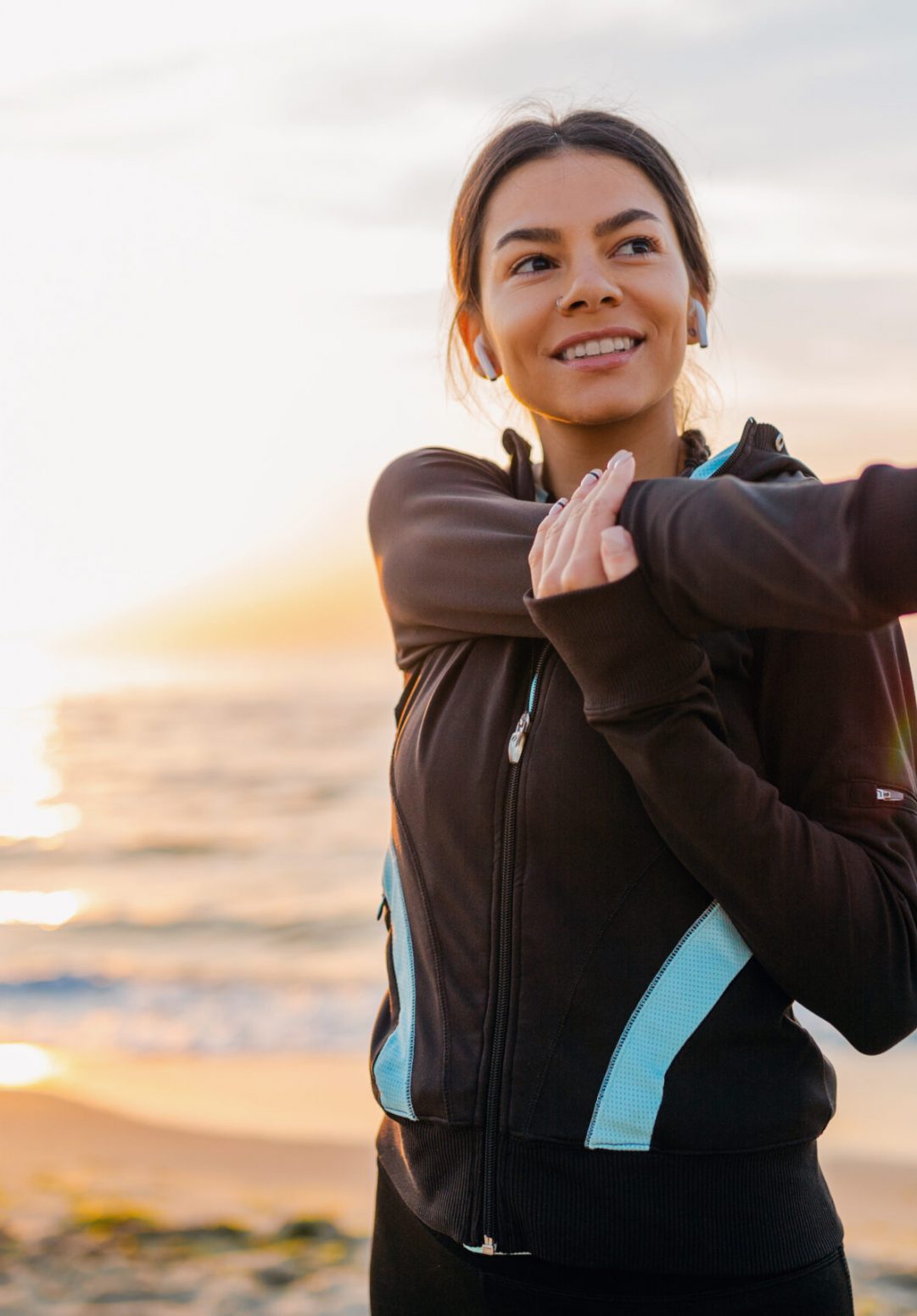 young attractive slim woman doing sport exercises on morning sunrise beach in sports wear, healthy lifestyle, listening to music on earphones, making stretching for arms
